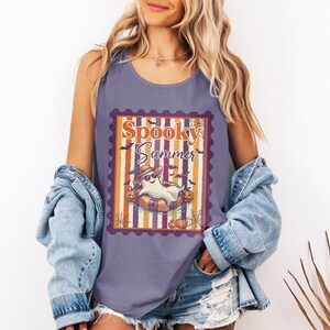 以下が含まれることがあります： 紫色のタンクトップ。前面には「Spooky Summer」のグラフィック。サングラスと帽子をかぶったお化けが浮き輪に乗っており、カボチャとコウモリが描かれています。柔らかい素材で作られています。