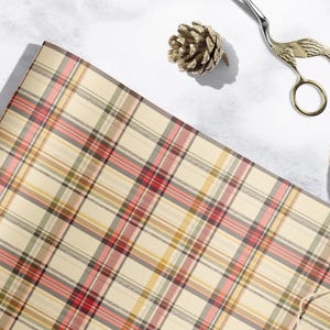 a plaid wrapping paper, a pair of scissors, a pine cone, and a spool of twine or string, all arranged on a white surface.