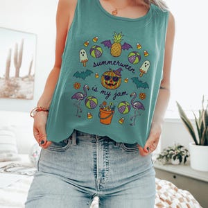 May include: Teal tank top with a circular graphic featuring a jack-o'-lantern wearing sunglasses, a pineapple with bat wings, and the text "Summerween is my jam." The design includes flamingos, candy corn, and beach balls.