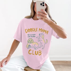 May include: A light pink t-shirt with the words "Doodle Mama Club" in gold lettering. The shirt features a graphic of two doodles, one holding a coffee cup. The text "Life is sweeter with Doodles" is also printed on the shirt.