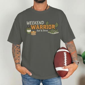 May include: A gray t-shirt with the words "WEEKEND WARRIOR" and the phrase "fuel & focus" printed on the front. The graphic includes a coffee cup, pancakes, a dumbbell, and sneakers. A brown football is held in the hands.