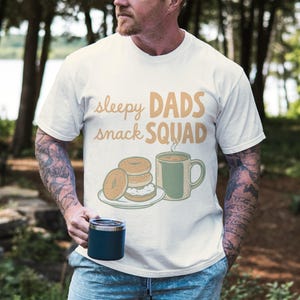 May include: A cream-coloured t-shirt with the text "sleepy DADS snack SQUAD" in brown and orange. The graphic features bagels, a mug of coffee, and a plate. The person in the image is holding a blue tumbler.