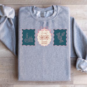 May include: A grey sweatshirt with rolled sleeves. The front displays three teal rectangular patches with science-themed illustrations and the text "SCIENCE BITCHES". The central patch features the formula E=mc².