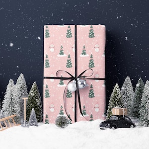A large pink gift box with a snowman and Christmas trees on it, surrounded by a snowy landscape with a black car and miniature trees.