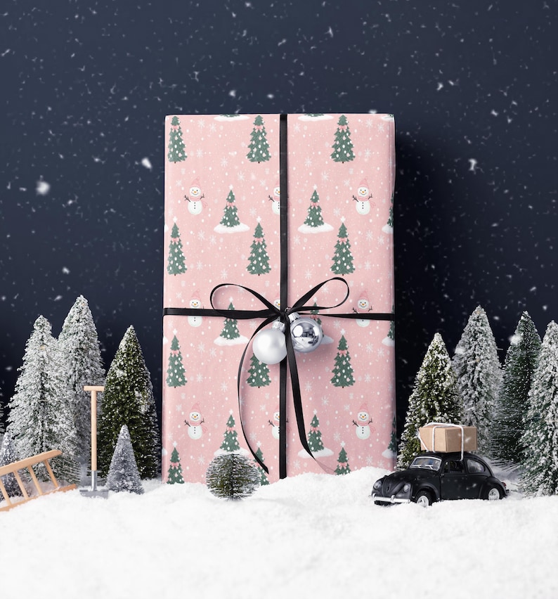 A large pink gift box with a snowman and Christmas trees on it, surrounded by a snowy landscape with a black car and miniature trees.
