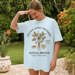 May include: Light blue t-shirt with a nature-themed graphic. The design features the text "CICADA SONG ORCHARD" and "ANNUAL BROODS" with illustrations of cicadas, leaves, and a teacup. The shirt is worn by a person outdoors.