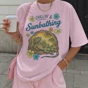 May include: Pink t-shirt with the text "Chillin' & Sunbathing" and a graphic of an iguana lounging in a cave. The shirt is paired with pink and white striped shorts. A gold chain necklace and bracelets complete the look.