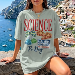 Op de afbeelding: Lichtgroen t-shirt met het woord "SCIENCE" in rode letters. Het ontwerp bevat een strik, bekers en een taartafbeelding. De tekst op het shirt luidt "LAB NERDS HOME & BRAVE" en "Pi Day of 3.14159".