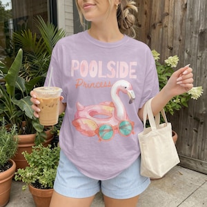 May include: A lilac t-shirt with the words "Poolside Princess" and a graphic of a flamingo float wearing sunglasses. The shirt is paired with blue and white striped shorts.