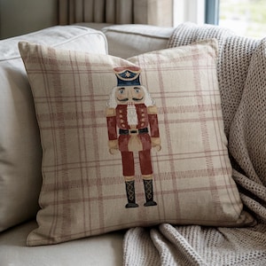 May include: Square decorative pillow with a plaid pattern in beige and red. The pillow features an embroidered image of a nutcracker soldier in red, black, and blue. The pillow is placed on a beige sofa with a knitted throw.