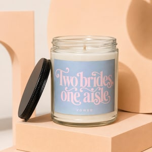 May include: A clear glass candle jar with a black metal lid resting beside it. The label is light blue with pink cursive text that reads Two brides, one aisle and the word VOWED in smaller white capital letters below the main text.