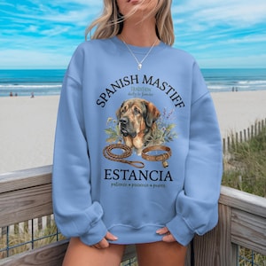 May include: A light blue sweatshirt featuring a watercolor illustration of a Spanish Mastiff dog. The design includes the text "Spanish Mastiff" and "Estancia" with the words "patience, presence, power". A leash and collar are also depicted.