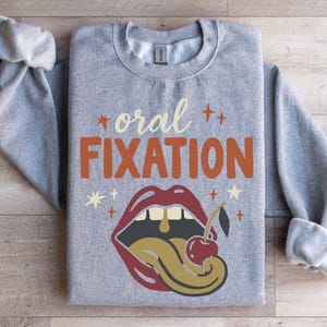 May include: A grey sweatshirt featuring the words "oral FIXATION" in orange and cream. The design includes red lips, a cherry, and a gold tongue. The sweatshirt has rolled-up sleeves.
