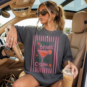 May include: A charcoal gray short-sleeve t-shirt featuring a graphic print of a pink martini glass with an orange slice. The text reads This Bride Needs a Cosmo, surrounded by pink vertical stripes and small star icons on a dark gray fabric background.