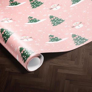 A roll of pink wrapping paper with a repeating pattern of Christmas trees and snowmen on it, placed on a wooden floor.
