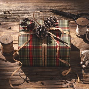A plaid gift box with pine cones and a pair of scissors sits on a wooden surface, surrounded by other holiday-themed items.