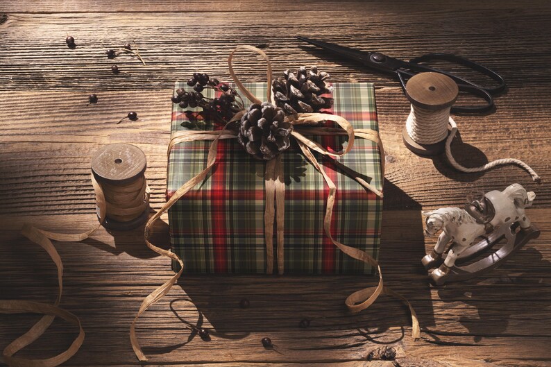 A plaid gift box with pine cones and a pair of scissors sits on a wooden surface, surrounded by other holiday-themed items.