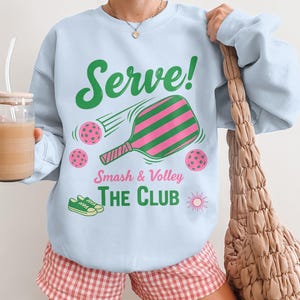 Könnte beinhalten: Hellblaues Sweatshirt mit dem Text "Serve!" und einer Grafik eines Pickleball-Schlägers und -Bälle. Die Worte "Smash & Volley The Club" sind ebenfalls auf dem Shirt aufgedruckt. Das Bild zeigt auch ein Paar grüne Turnschuhe und eine Sonne.