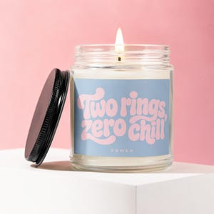 May include: A clear glass candle jar with a lit wick and a black lid resting beside it. The label is light blue with pink text that reads Two rings, zero chill and VOWED in a smaller font. The candle contains white wax and emits a flame.