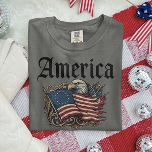 May include: A gray Comfort Colors t-shirt featuring the word "America" in a gothic font. The shirt displays an illustration of an eagle, American flags, and decorative scrollwork. The shirt is laid on a red and white striped surface.