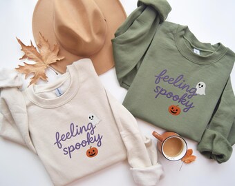 May include: Two sweatshirts with the text "feeling spooky" in purple script, a ghost, and a jack-o'-lantern. One sweatshirt is cream-colored, the other is olive green. A tan hat and a fall leaf are also in the image.