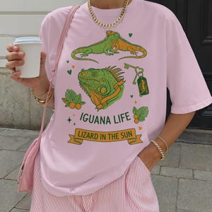Op de afbeelding: Een lichtroze t-shirt met een groene en oranje leguaan-afbeelding en de tekst "IGUANA LIFE LIZARD IN THE SUN". De shirt heeft ook een sleutel en illustraties van sinaasappels. De persoon draagt roze en wit gestreepte shorts.