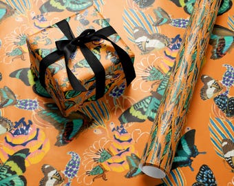 Puede incluir: Caja de regalo naranja envuelta en papel con estampado de mariposas, atada con una cinta de raso negra. También se ve un rollo de papel de regalo a juego. El diseño de mariposas presenta una variedad de colores, incluyendo azul, verde y amarillo.