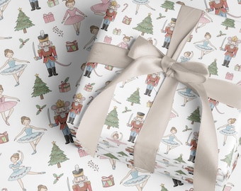 May include: White wrapping paper with a repeating pattern of ballerinas, nutcrackers, Christmas trees, and gift boxes. A beige ribbon is tied around the wrapped package. The design has a whimsical, festive feel.