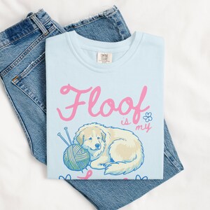 May include: A light blue t-shirt with the text "Floof is my love language" in pink script. The shirt features a cartoon dog with a ball of yarn and knitting needles. The shirt is laid on top of a pair of blue jeans.
