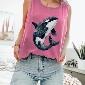Könnte beinhalten: Rosafarbenes Tanktop mit einer Aquarellillustration eines schwarz-weißen Orca-Wals. Das ärmellose Oberteil ist aus einem weichen Material gefertigt und eignet sich für Freizeitkleidung. Das Orca-Wal-Design ist mittig auf der Vorderseite des Shirts platziert.