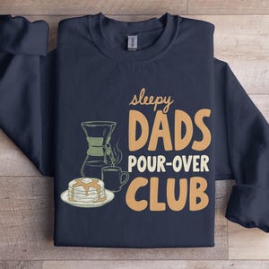May include: Navy blue sweatshirt featuring the text "sleepy DADS POUR-OVER CLUB" in orange and white. The design includes illustrations of a coffee pour-over, pancakes, and a mug. The sweatshirt is folded on a wooden surface.
