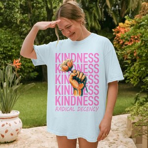 Könnte beinhalten: Hellblaues T-Shirt mit dem Wort "KINDNESS" in Rosa, vertikal wiederholt. Eine Grafik einer Hand, die eine Blume hält, befindet sich in der Mitte. Die Worte "RADICAL DECENCY" sind unten auf dem T-Shirt aufgedruckt.