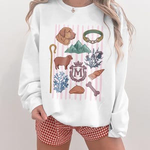 Op de afbeelding: Witte sweatshirt met illustraties van een hondenhoofd, bergen, een schaap, een herdersstaf en een bot. Het ontwerp is geplaatst tegen een achtergrond van roze en witte verticale strepen.