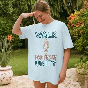 Può includere: T-shirt azzurra con le parole "WALK FOR PEACE UNITY" in verde acqua e rosso. La maglietta presenta una grafica di una persona con un simbolo di pace. La persona indossa una veste e cammina.