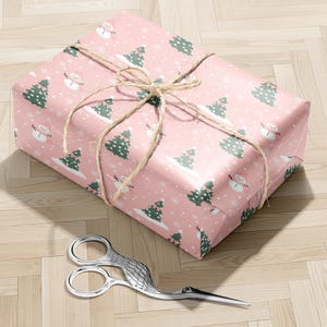 A pink gift box with a snowman and Christmas tree pattern wrapped in twine, placed on a wooden floor next to a pair of scissors.