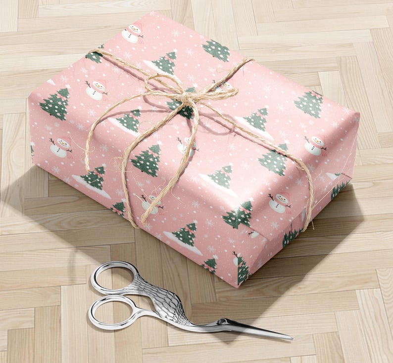 A pink gift box with a snowman and Christmas tree pattern wrapped in twine, placed on a wooden floor next to a pair of scissors.