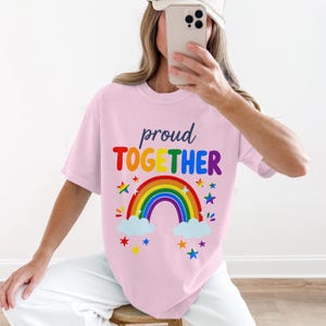 Puede incluir: Camiseta rosa claro con las palabras "proud TOGETHER" en colores arcoíris. Un arcoíris con nubes y estrellas está debajo del texto. La camiseta es de estilo casual.