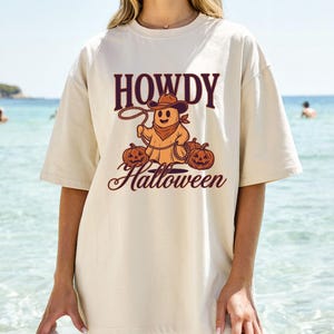 Op de afbeelding: Crèmekleurig T-shirt met de tekst "HOWDY Halloween" in een western-thema. De afbeelding toont een spook met een cowboyhoed en lasso, met twee uitgesneden pompoenen eronder.