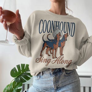 May include: Beige sweatshirt with a graphic of two Coonhound dogs howling at the moon. The text "COONHOUND" is arched above the dogs, with "Sing Along" in a script font below. The sweatshirt has long sleeves.