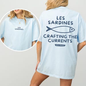 以下が含まれることがあります： 薄い青色のTシャツに、魚のグラフィックと「Les Sardines Crafting the Currents Sete, France」というテキストがプリントされています。