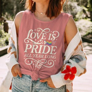Puede incluir: Una camiseta sin mangas rosa polvoriento con las palabras "LOVE IS PRIDE ALL YEAR LONG" en escritura blanca. El diseño incluye una bandera arcoíris y símbolos de género. La parte superior está anudada en la cintura. La persona lleva una rebeca blanca y beige y vaqueros azules.