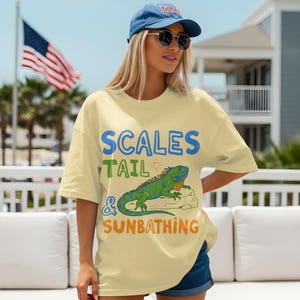 May include: A light yellow t-shirt with the text "SCALES TAIL & SUNBATHING" and a graphic of a green and blue iguana. The person is wearing a blue baseball cap and sunglasses.