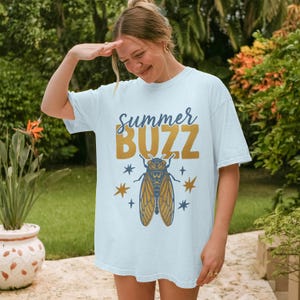 Puede incluir: Camiseta azul claro con la frase "Summer Buzz" en dorado y azul. Debajo, una ilustración detallada de una cigarra con estrellas. La camiseta tiene un corte holgado y casual.