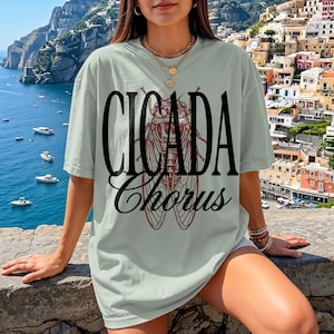 May include: A light green t-shirt with the words "CICADA Chorus" in black and red. The design features a detailed illustration of a cicada insect. The shirt is a relaxed fit.