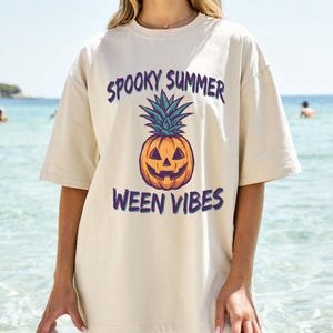 Può includere: T-shirt beige con la scritta "Spooky Summer Ween Vibes" in viola. La maglietta presenta una grafica di ananas-zucca arancione, verde e viola. Indossata in una giornata di sole in spiaggia.