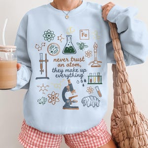 Peut inclure: Sweat-shirt bleu clair orné d'illustrations scientifiques et de la phrase « never trust an atom, they make up everything ». Le motif comprend des atomes, un microscope et du matériel de laboratoire. Un style de sweat-shirt décontracté et confortable.