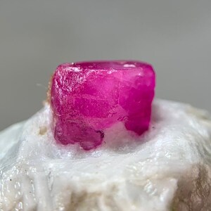 May include: Close-up of a vibrant, raw ruby gemstone. The ruby, a deep magenta hue, is set against a backdrop of white and green mineral. The rough texture of the stone is visible, highlighting its natural form. A specimen of raw mineral.