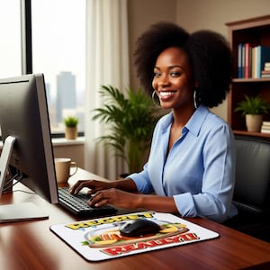 May include: A woman in a light blue button-down shirt smiles while working at a desk. A computer monitor, keyboard, and mouse are visible. A mousepad with the words "Regret is Really!" is in front of the mouse.