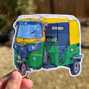 May include: A sticker featuring a colorful illustration of an auto rickshaw, with a yellow roof, green body, and blue accents. The sticker has the text "HYDERABADDIE" and a license plate number. The background is blurred.