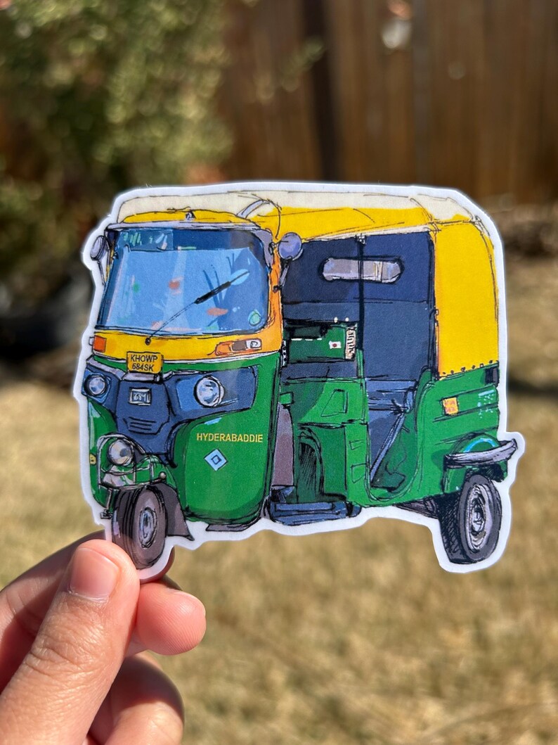 May include: A sticker featuring a colorful illustration of an auto rickshaw, with a yellow roof, green body, and blue accents. The sticker has the text "HYDERABADDIE" and a license plate number. The background is blurred.